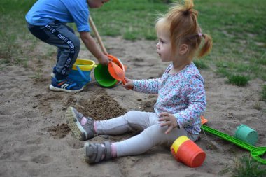 Smiling toddler girl playing with sand in a sandbox, holding a small orange shovel while her brother digs nearby with colorful buckets and toys, enjoying playful outdoor activity.