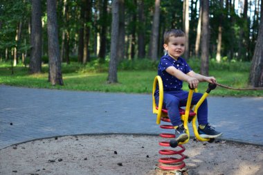 Smiling boy playing on spring rocker at playground, holding stick while enjoying outdoor summer fun in park. Happy child in blue clothes having leisure time.