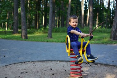 Smiling boy playing on spring rocker at playground, holding stick while enjoying outdoor summer fun in park. Happy child in blue clothes having leisure time.