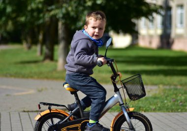 Focused little boy riding orange bike with basket in city park. Toddler in grey hoodie and blue scarf learning to cycle, enjoying outdoor play, sport, and family activity on a bright day.