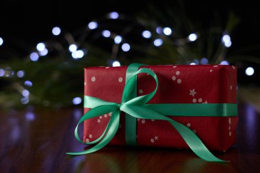 A gift box with a ribbon placed on a brown table, set against a backdrop of a pine branch decorated with twinkling fairy lights.