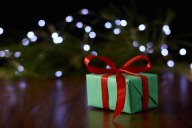 A gift box with a ribbon placed on a brown table, set against a backdrop of a pine branch decorated with twinkling fairy lights.