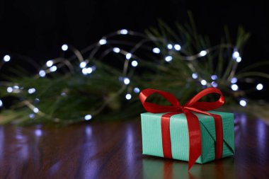 A gift box with a ribbon placed on a brown table, set against a backdrop of a pine branch decorated with twinkling fairy lights.