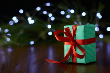 A gift box with a ribbon placed on a brown table, set against a backdrop of a pine branch decorated with twinkling fairy lights.