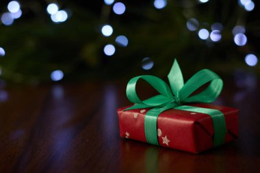 A gift box with a ribbon placed on a brown table, set against a backdrop of a pine branch decorated with twinkling fairy lights.