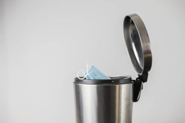 Açık çöp tenekesinin içinde tek kullanımlık mavi yüz maskesi temiz gri arkaplandan bakıldığında hijyen ve uygun atık imhası vurgulanıyor