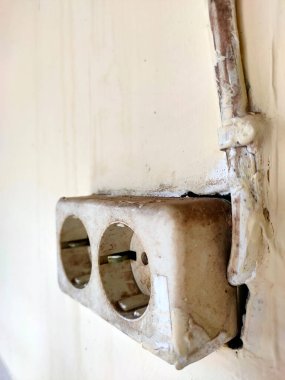 Kirli, yaşlanmış çift prizli bir prizin yakın çekimi. Duvara monte edilmiş. Bu yıpranmış durum, zamanın geçişini ve potansiyel elektrik güvenliği endişelerini vurguluyor..