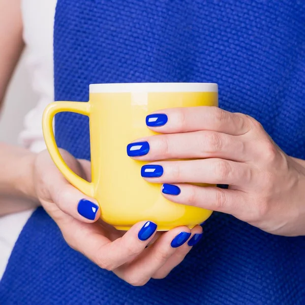 Vackra kvinnliga händer som håller gul kopp. Manikyr med blått nagellack. Mysigt koncept — Stockfoto