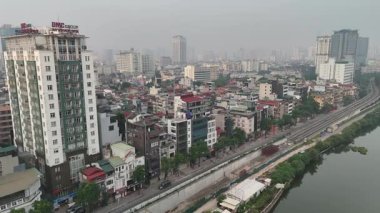Hanoi, Vietnam 'daki Thu Le Lake' in 4K hava görüntüsü, su, yürüyüş yolları ve şehrin yüksek ufuk çizgisinin arka planında yer alan ağaçla kaplı huzurlu yeşil bir vahayı gözler önüne seriyor. Bu manzaralı kentsel göl,