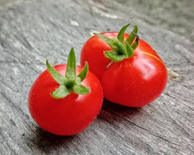 Yeşil saplı iki taze kırmızı domates yıpranmış ahşap bir yüzeyde dinleniyor ve kırsal mutfak cazibesini çağrıştırıyor. Parlak, sağlıklı ürünler salatalar, atıştırmalıklar ve renkli yemek fotoğrafçılığı için idealdir..
