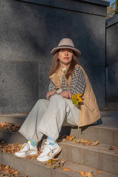 Bir kadın, düşen sonbahar yapraklarıyla çevrili taş basamaklarda rahatlar. Sarı bir yaprak tutuyor ve şık bir kıyafet giyiyor. Parkta güneşli bir günün tadını çıkarıyor..