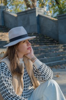 Bir kadın parkta taş basamaklarda oturuyor, düşüncelere dalmış. Şık bir şapka ve rahat bir süveter giyer. Düşen yaprakların arasında sıcak güneş ışığının tadını çıkarır..