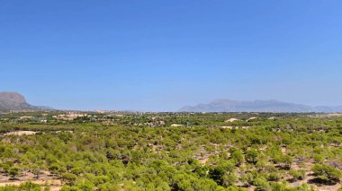 Berrak bir gökyüzünün altındaki yemyeşil ve dağlık araziyi gösteren nefes kesici panoramik bir manzara.