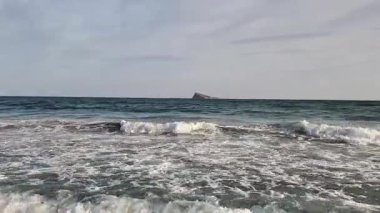 Huzurlu ve huzurlu bir okyanus manzarası bulutlu bir gökyüzünün altındaki kayalık bir oluşumda hafif dalgalar sergiliyor.