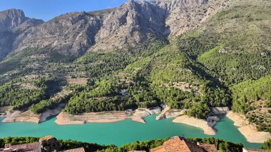 Çarpıcı manzara, açık mavi gökyüzünün altında turkuaz bir su deposunu çevreleyen yeşil dağları sergiliyor.