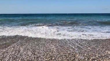 Yumuşak dalgalar, yukarıdaki berrak ve canlı mavi gökyüzünün altında, resmedilmeye değer bir çakıl taşı plajına hafifçe çarpıyor.