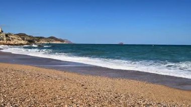 Güzel bir sahil sahnesi, yumuşak dalgalar, çakıllı kıyılar ve berrak bir gökyüzünün altında sakin bir atmosfer..