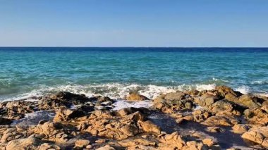 Güzel ve huzurlu bir dalga manzarası yukarıdaki açık mavi gökyüzünün altında kayalık bir plaja usulca çarpıyor.
