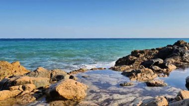 Güzel ve sakin bir kıyı manzarası berrak mavi suları ve parlak gökyüzünün altındaki engebeli kayaları sergiliyor.