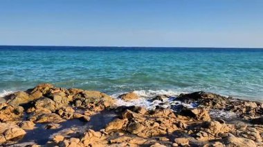 Stunning ocean waves continuously crash powerfully against the rugged rocks beneath a clear blue expanse of sky