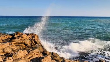 Güzel okyanus dalgaları zarif bir şekilde kayalık bir kıyı şeridine çarparken gökyüzü mükemmel bir şekilde berrak ve parlak kalır.