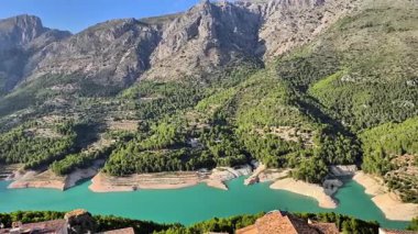 Çarpıcı manzara, açık mavi gökyüzünün altında turkuaz bir su deposunu çevreleyen yeşil dağları sergiliyor.