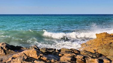 Okyanus dalgalarının, berrak mavi gökyüzünün altındaki engebeli kayalara çarpması nefes kesici bir şekilde büyüleyici bir manzara.
