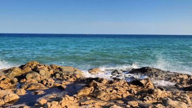 Stunning ocean waves continuously crash powerfully against the rugged rocks beneath a clear blue expanse of sky