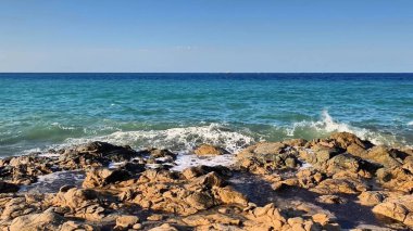 Güzel ve huzurlu bir dalga manzarası yukarıdaki açık mavi gökyüzünün altında kayalık bir plaja usulca çarpıyor.