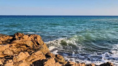 A beautifully serene view of ocean waves gently interacting with rugged rocks beneath a clear blue sky