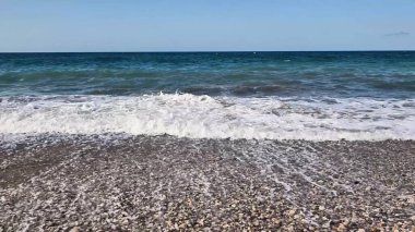 Yumuşak dalgalar, yukarıdaki berrak ve canlı mavi gökyüzünün altında, resmedilmeye değer bir çakıl taşı plajına hafifçe çarpıyor.