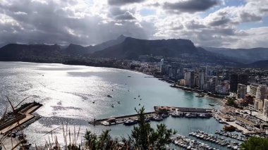 Experience a stunning panoramic view of a vibrant coastal city, with a marina, majestic mountains, and tranquil water