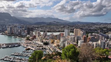 This aerial view highlights a vibrant coastal city, with a marina and majestic mountains under a stunning sky