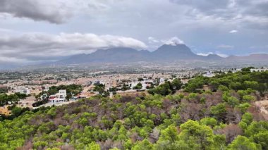A stunning view of majestic mountains, a vibrant city, and lush greenery under a captivating cloudy sky