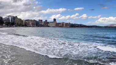 Güzel bir kıyı şeridi sahnesi, açık mavi gökyüzünün altında modern binalarla kıyıya vuran hafif dalgaları gösteriyor.