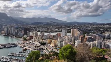 This aerial view highlights a vibrant coastal city, with a marina and majestic mountains under a stunning sky