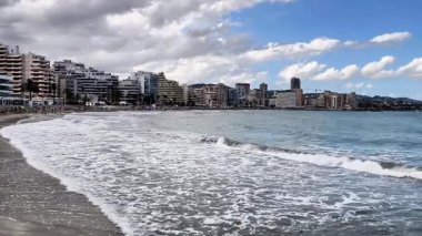 Sahile vuran dalgalarla dolu bir sahil sahnesi, çarpıcı bir şehir silüeti, ve güzel bulutlu bir gökyüzü.