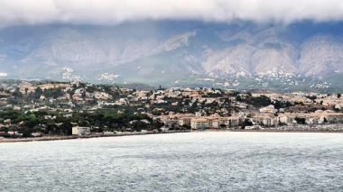 A beautiful coastal view of a vibrant city by the sea, with majestic mountains and a cloudy sky above