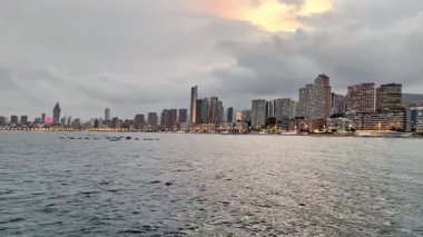 Gün batımında güzel bir kıyı şehrinin ufuk çizgisi aşağıdaki sakin suların üzerinde ışıldayan parlak ışıkları yansıtır.