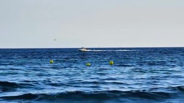 Hız yapan bir teknesi, renkli şamandıraları ve açık, güneşli bir gökyüzünün altında hafif dalgaları olan sakin bir okyanus manzarası.