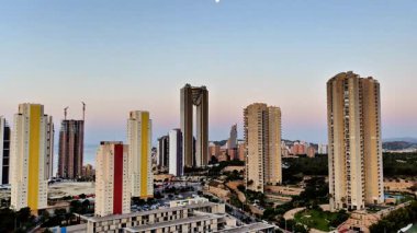 Kentsel ufuk çizgisi, açık mavi gökyüzünün zeminine kurulmuş çeşitli yüksek binalar sergiliyor.