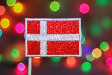 Havai fişek ve bayram kutlamalarına benzeyen şenlikli bokeh ışıklarının arka planıyla aydınlatılmış nakışlı bir Danimarka bayrağı. Noel, Yeni Yıl ve ulusal gurur teması için ideal.