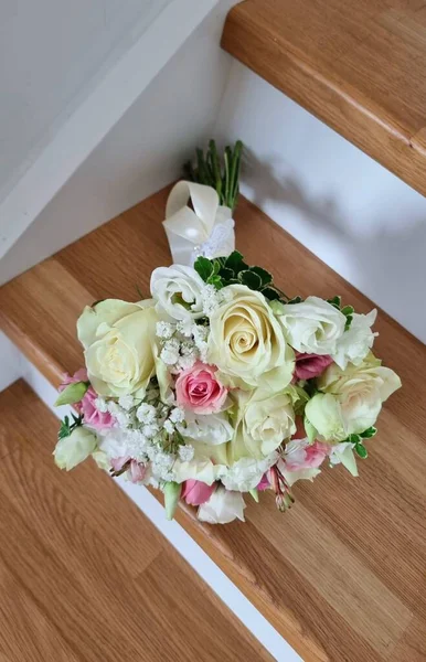 Gül, östoma, çingene ve yeşillikten yapılmış pembe ve beyaz renkli yuvarlak bir düğün buketi. Buket kendi sapında, tahta merdivenlerde..