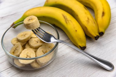 Çiğ sarı muz meyve bir kase katiyen Beyaz ahşap masa üzerinde bir çatalı dilimler.