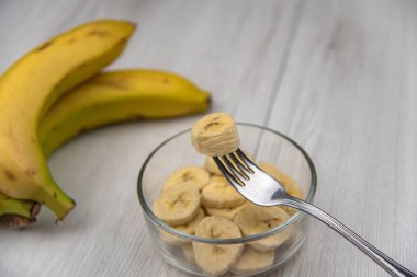 Çiğ sarı muz meyve bir kase katiyen Beyaz ahşap masa üzerinde bir çatalı dilimler.