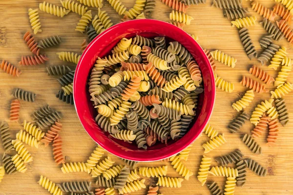 stock image Raw uncooked three-colored Fusilli durum wheat pasta with spinach and tomato in a red bowl on wood table, top view.