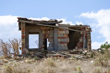 Parçalanmış bulutlu bir gökyüzünün altında dağınık enkaz ve seyrek bitki örtüsüyle çevrili, kurumuş bir arazide terk edilmiş tuğla bir yapı duruyor..