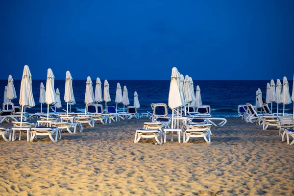 Düzgün bir şekilde düzenlenmiş beyaz şezlonglar ve şemsiyeler sakin bir alacakaranlık sahili boyunca uzanan derin mavi bir ufuk çizgisi..
