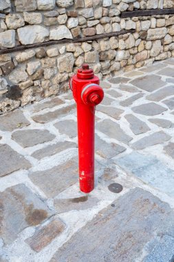 Parlak kırmızı bir yangın musluğu engebeli bir taş kaldırımda duruyor sakin bir kentin köşesinde yıpranmış bir taş duvara karşı..