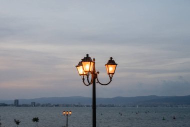 Klasik tarzda bir sokak lambası, kıyı kenarında alacakaranlıkta sıcacık parlıyor. Kasvetli bir gökyüzünün altındaki uzak tepeler ve binalar....
